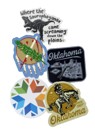 A group of colorful die-cut stickers arranged on a wooden surface. The stickers feature various designs including a black dinosaur with the text 'Where the saurophaganax came screaming down the plains', a blue circular design with a green pine branch and white feathers, a dark blue 'Oklahoma' sticker with a bird and landscape, a white and blue geometric snowflake design, and a gold-toned 'Oklahoma Since 1907' sticker with a cowboy on a horse.